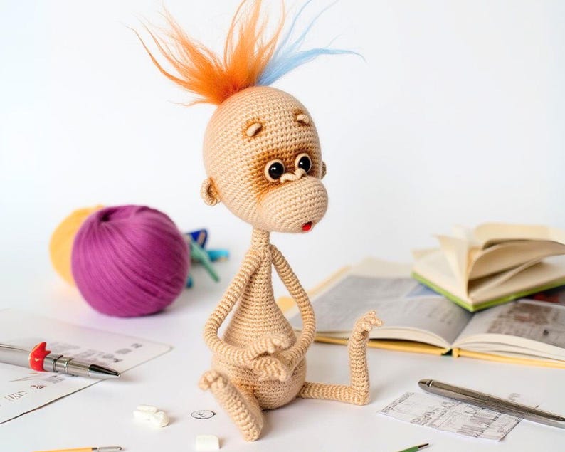 May include: A crocheted monkey toy with a brown face, orange and blue yarn hair, and a red mouth. The monkey is sitting on a white surface with yarn balls and books in the background.
