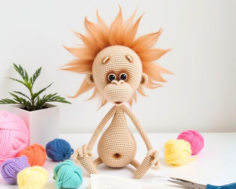 May include: A crocheted stuffed monkey toy with a light brown body, brown eyes, and a fluffy orange mane. The monkey is sitting on a white surface with colorful yarn balls.
