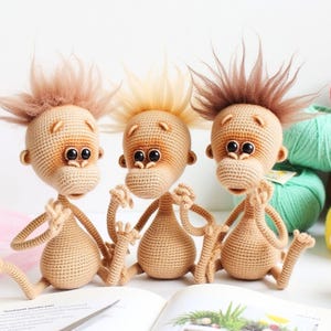 May include: Three crocheted monkey plushies with brown fur, sitting on a book. The monkeys have brown eyes and are wearing a neutral expression.