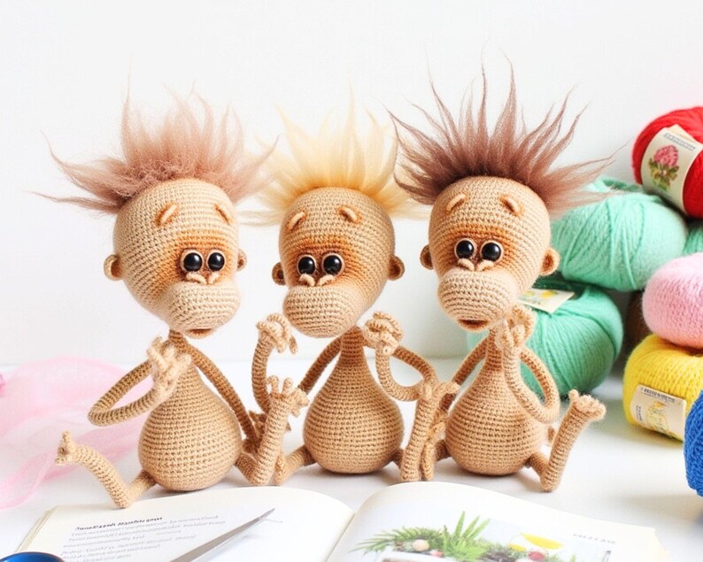 May include: Three crocheted monkey plushies with brown fur, sitting on a book. The monkeys have brown eyes and are wearing a neutral expression.