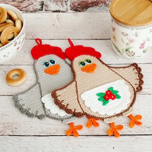以下が含まれることがあります： 鶏の形をした、かぎ針編みで作った鍋つかみが2つ。1つは赤色の帽子をかぶったグレー、もう1つは白いエプロンと赤い実をつけたブラウンです。
