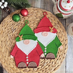 Könnte beinhalten: Zwei handgefertigte Topflappen in Form von gehäkelten Zwergen. Einer hat einen roten Hut und einen grünen Körper, der andere hat einen grünen Hut und einen roten Körper. Beide haben weiße Bärte, rosa Nasen und braune Schuhe. Sie liegen auf einem geflochtenen Platzset mit Weihnachtsschmuck.