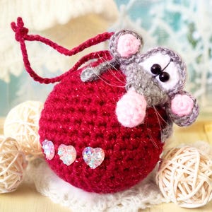 May include: A red crocheted Christmas ornament with a gray crocheted mouse attached to it. The ornament has sparkly pink hearts on it.
