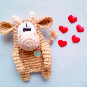 May include: A crocheted brown cow with a pink nose and big black eyes. The cow is surrounded by red hearts.