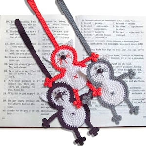 May include: Three crocheted cat bookmarks in red, gray, and black. Each bookmark has a white face with black eyes and a pink nose. The bookmarks are attached to a book.