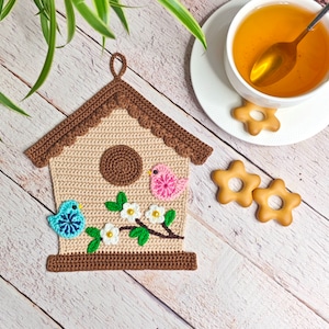 Puede incluir: Un posavasos de ganchillo marrón en forma de casita de pájaros con un pájaro rosa y azul, flores blancas y hojas verdes.