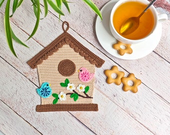 May include: A crocheted brown birdhouse pot holder with a pink and blue bird, white flowers, and green leaves.