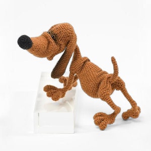 Może przedstawiać: Pluszowa zabawka w kształcie brązowego psa z długim pyskiem i czarnym nosem. Pies stoi na tylnych łapach, a przednie łapy opiera o biały blok.