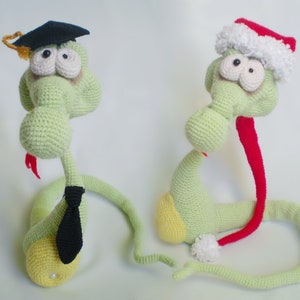 Könnte beinhalten: Zwei gehäkelte grüne Schlangenspielzeuge, eines mit einer schwarzen Abschluss-Kappe und einer Krawatte, das andere mit einer rot-weißen Weihnachtsmütze. Beide haben große Glitz Augen und gelbe Bäuche.