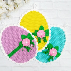 Könnte beinhalten: Drei gehäkelte Topflappen in Form von Ostereiern in Rosa, Gelb und Blau mit weißem Rand. Jeder Topflappen hat eine grüne gehäkelte Blume mit einem rosa Zentrum.