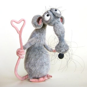 Pode incluir: Um brinquedo de rato de feltro cinza segurando um pau rosa em forma de cora&ccedil;&atilde;o. O rato tem olhos pretos grandes e uma cauda longa e cinza.