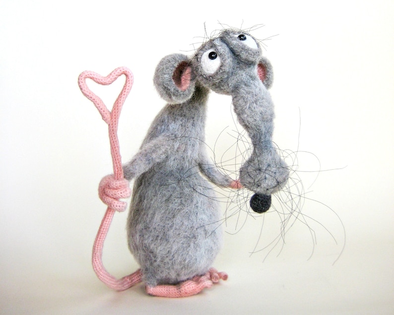 Pode incluir: Um brinquedo de rato de feltro cinza segurando um pau rosa em forma de cora&ccedil;&atilde;o. O rato tem olhos pretos grandes e uma cauda longa e cinza.