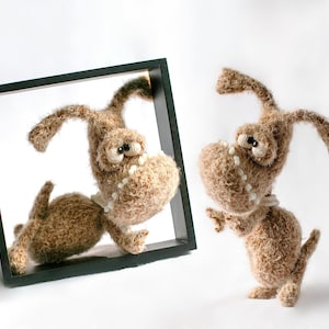 May include: A brown crocheted dog toy with a toothy grin is looking out of a black picture frame. Another brown crocheted dog toy with a toothy grin is standing to the right of the frame.