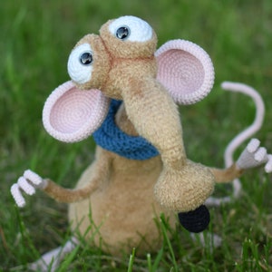 Pode incluir: Um brinquedo de pel&uacute;cia de rato de croch&ecirc; com olhos grandes, um focinho longo e uma echarpe azul. O rato est&aacute; sentado em um leito de grama verde.