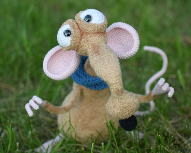 Pode incluir: Um brinquedo de pel&uacute;cia de rato de croch&ecirc; com olhos grandes, um focinho longo e uma echarpe azul. O rato est&aacute; sentado em um leito de grama verde.