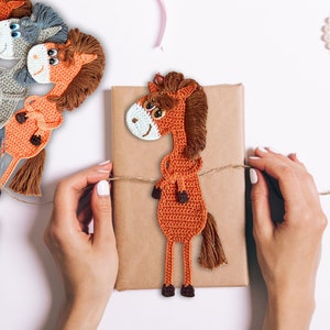 Può includere: Un segnalibro a forma di cavallo in crochet marrone con occhi bianchi e una criniera marrone. Il segnalibro è legato a una scatola regalo marrone con uno spago.