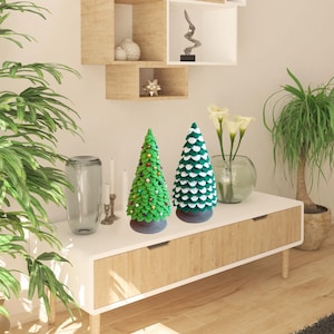 May include: Two green and white felt Christmas trees on a wooden shelf with a white top. The tree on the left has red and gold ornaments. The tree on the right has white accents.