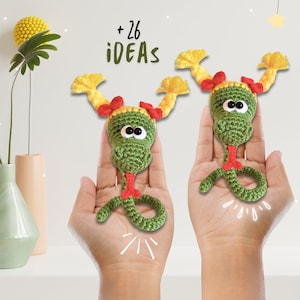 Puede incluir: Dos juguetes de serpiente de crochet verde con pelo de lana amarillo y lazos rojos. Las serpientes están sostenidas en las manos de dos personas. El texto + 26 IDEAS está por encima de las serpientes.