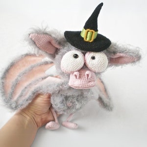 May include: A gray and pink crocheted bat wearing a black witch hat with a green band. The bat has large white eyes and a pink nose.