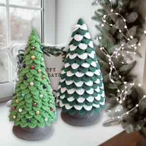 May include: Two crocheted Christmas trees, one green with colorful ornaments and one white and green, sitting on a windowsill with a white pillow behind them that says "Happy Days".