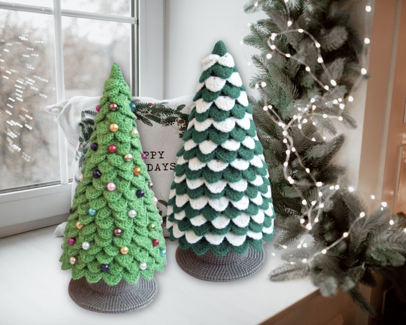 May include: Two crocheted Christmas trees, one green with colorful ornaments and one white and green, sitting on a windowsill with a white pillow behind them that says "Happy Days".