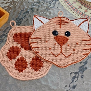 May include: Two crocheted cat-shaped pot holders. One pot holder is a brown paw print and the other is a light brown cat face with black eyes and a brown nose.