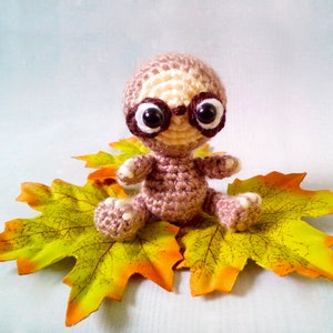 May include: A brown and white crocheted sloth wearing glasses sits on a bed of yellow and orange autumn leaves.