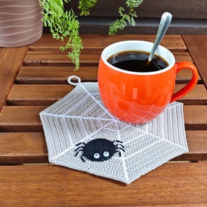 May include: A crocheted spiderweb coaster with a black spider in the center. The coaster is on a wooden table with a cup of coffee and a spoon.
