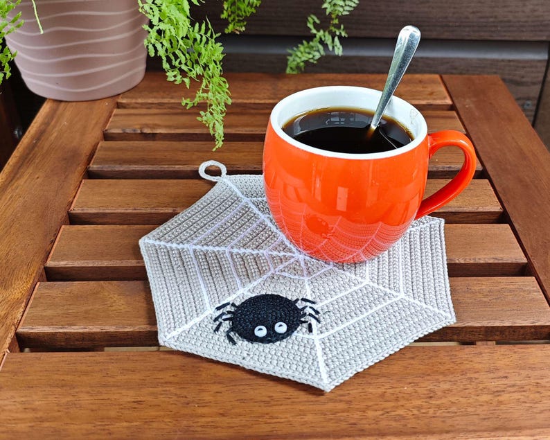 May include: A crocheted spiderweb coaster with a black spider in the center. The coaster is on a wooden table with a cup of coffee and a spoon.