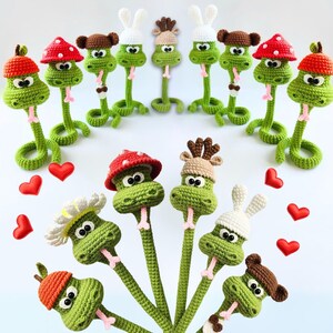 Puede incluir: Una colección de juguetes de serpiente de crochet con varios sombreros, incluyendo un hongo, una flor y un conejito. Las serpientes son verdes y tienen lenguas rosadas. Los juguetes están dispuestos en círculo sobre un fondo blanco.