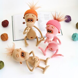 May include: Three crocheted monkey toys with different colored hats. The monkeys are beige, pink, and orange. The monkeys are sitting on a book and a white surface.