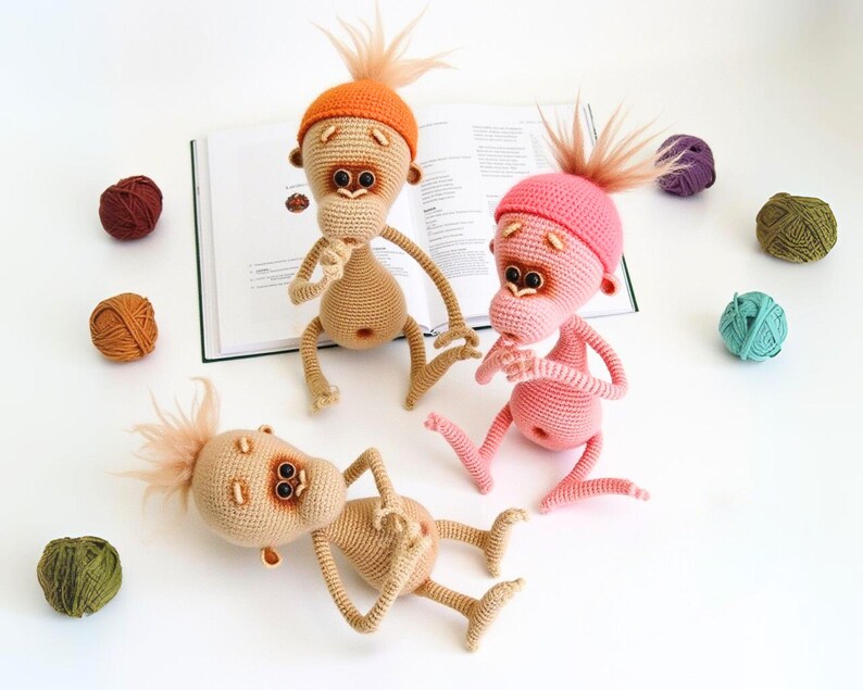 May include: Three crocheted monkey toys with different colored hats. The monkeys are beige, pink, and orange. The monkeys are sitting on a book and a white surface.