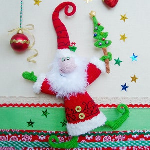 May include: A crocheted Santa Claus doll with a white beard and a red hat, holding a small green Christmas tree. The doll is standing on a green and red striped background with white stars.