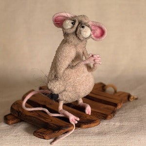 Pode incluir: Um rato cinzento de croch&ecirc; com orelhas cor-de-rosa e olhos grandes est&aacute; sentado em uma plataforma de madeira. O rato tem uma cauda longa e &eacute; feito com um fio felpudo.