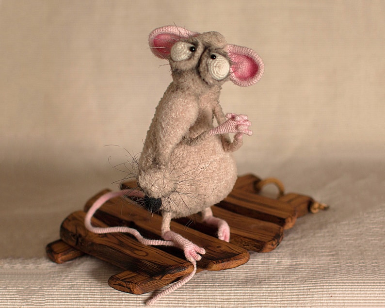 Pode incluir: Um rato cinzento de croch&ecirc; com orelhas cor-de-rosa e olhos grandes est&aacute; sentado em uma plataforma de madeira. O rato tem uma cauda longa e &eacute; feito com um fio felpudo.
