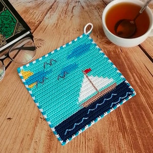 Könnte beinhalten: Gehäkelter quadratischer Topflappen mit einem Segelboot-Design, das eine rote Flagge vor einem blau-weißen Himmel und Meer zeigt. Der Topflappen hat einen weißen Rand und eine Schlaufe zum Aufhängen. Eine Tasse Tee und ein Buch sind im Hintergrund.