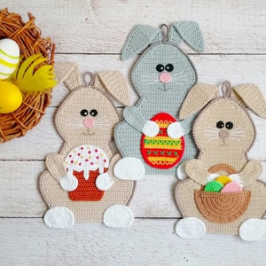 May include: Crocheted Easter bunny decorations in beige and gray, each with a unique design. One bunny holds a cake, another an Easter egg, and the third a basket of eggs. A woven nest with decorated eggs is in the background.