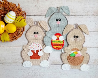 May include: Crocheted Easter bunny decorations in beige and gray, each with a unique design. One bunny holds a cake, another an Easter egg, and the third a basket of eggs. A woven nest with decorated eggs is in the background.