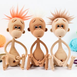 May include: Three crocheted monkey toys with different colored hair. The monkey on the left has orange hair, the middle monkey has white hair, and the monkey on the right has brown hair. All three monkeys are sitting on a white surface.