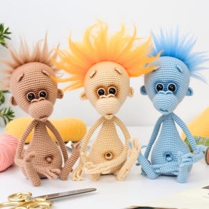 May include: Three crocheted monkey toys sitting on a white surface. The monkey on the left has brown fur and a brown mohawk. The monkey in the middle has light brown fur and a bright orange mohawk. The monkey on the right has blue fur and a light blue mohawk.