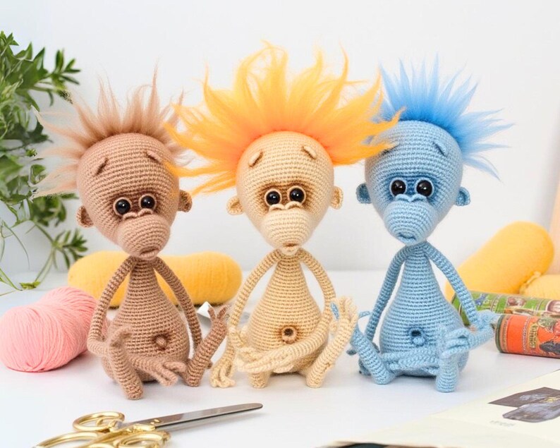 May include: Three crocheted monkey toys sitting on a white surface. The monkey on the left has brown fur and a brown mohawk. The monkey in the middle has light brown fur and a bright orange mohawk. The monkey on the right has blue fur and a light blue mohawk.