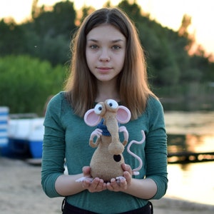 Pode incluir: Uma pessoa jovem segurando um brinquedo de rato de croch&ecirc; marrom e rosa com olhos grandes e uma echarpe azul. O brinquedo tem um nariz longo e fino e um corpo pequeno e redondo.