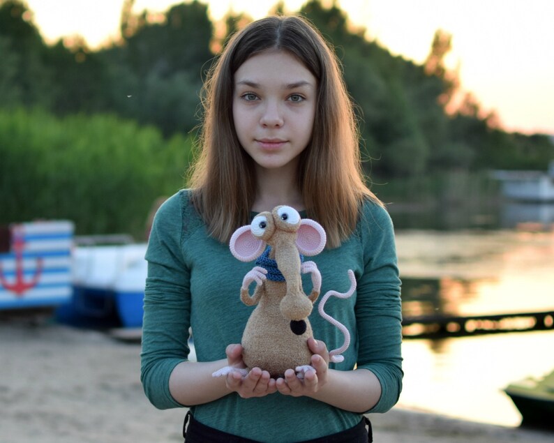 Pode incluir: Uma pessoa jovem segurando um brinquedo de rato de croch&ecirc; marrom e rosa com olhos grandes e uma echarpe azul. O brinquedo tem um nariz longo e fino e um corpo pequeno e redondo.