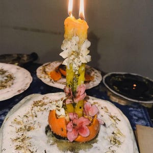 Puede incluir: Una vela de pilar amarilla con dos mechas encendidas, incrustada con p&eacute;talos de flores secas blancas y rosas. La vela est&aacute; colocada dentro de una c&aacute;scara de naranja ahuecada, sobre un plato blanco cubierto con una capa de hierbas secas.