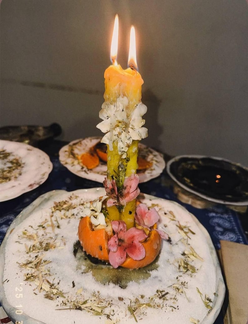 Puede incluir: Una vela de pilar amarilla con dos mechas encendidas, incrustada con p&eacute;talos de flores secas blancas y rosas. La vela est&aacute; colocada dentro de una c&aacute;scara de naranja ahuecada, sobre un plato blanco cubierto con una capa de hierbas secas.