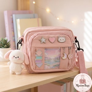 May include: A pink crossbody bag with multiple pockets and a matching strap. The bag features decorative pins, including a star, heart, and cat face. A plush bunny keychain hangs from the side. The bag is displayed on a wooden shelf.