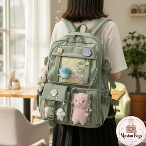Puede incluir: Una mochila de color verde salvia con un bolsillo frontal transparente y un fondo amarillo con estampado de nubes. La bolsa está decorada con varios colgantes de peluche, incluyendo una llama rosa, un conejo blanco y una criatura azul.