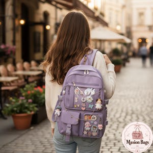 Könnte beinhalten: Ein hellvioletter Rucksack mit einer durchsichtigen Vordertasche, die mit verschiedenen kleinen dekorativen Anstecknadeln und Anhängern gefüllt ist. Der Rucksack verfügt über mehrere Reißverschlussfächer, eine Schnallentasche und Anhänger.