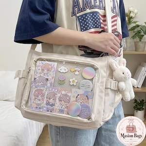 Puede incluir: Bolso mensajero beige con un bolsillo frontal transparente lleno de pegatinas y pines. Un conejo de peluche blanco cuelga del lateral. El bolso tiene una correa para el hombro y un pequeño bolsillo lateral.