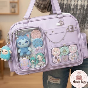 May include: A light purple ita bag featuring two clear display windows filled with various circular pins, a small blue plush cat, and two small anime-style character figures. A fluffy blue sheep keychain hangs from the side, and a silver chain decorates the front.
